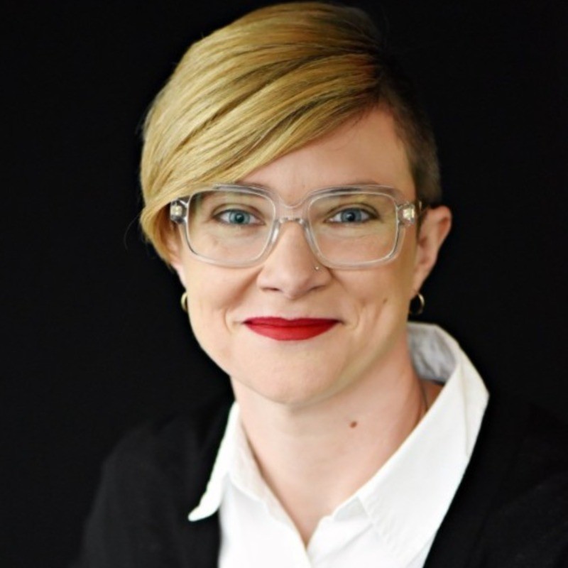 Professional headshot of a person with short blond hair, clear glasses, red lipstick, and black blazer on dark background.