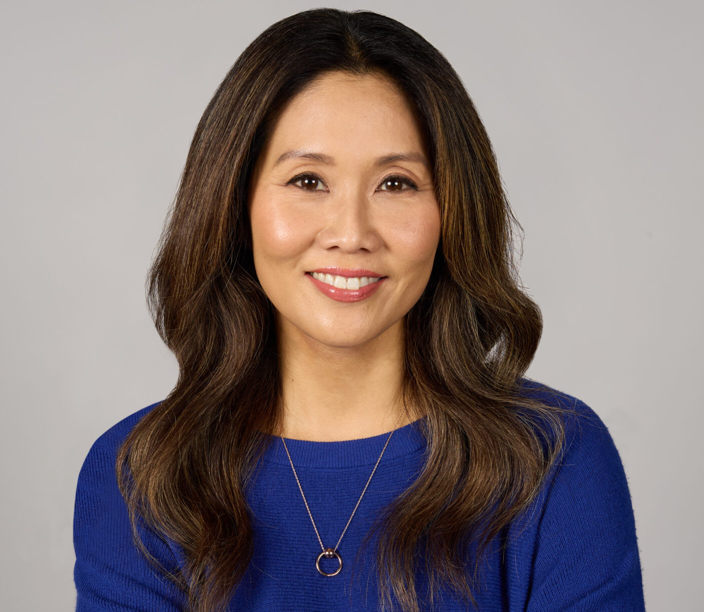 Professional headshot of a woman with long dark hair, blue sweater, necklace, and crossed arms on neutral background.