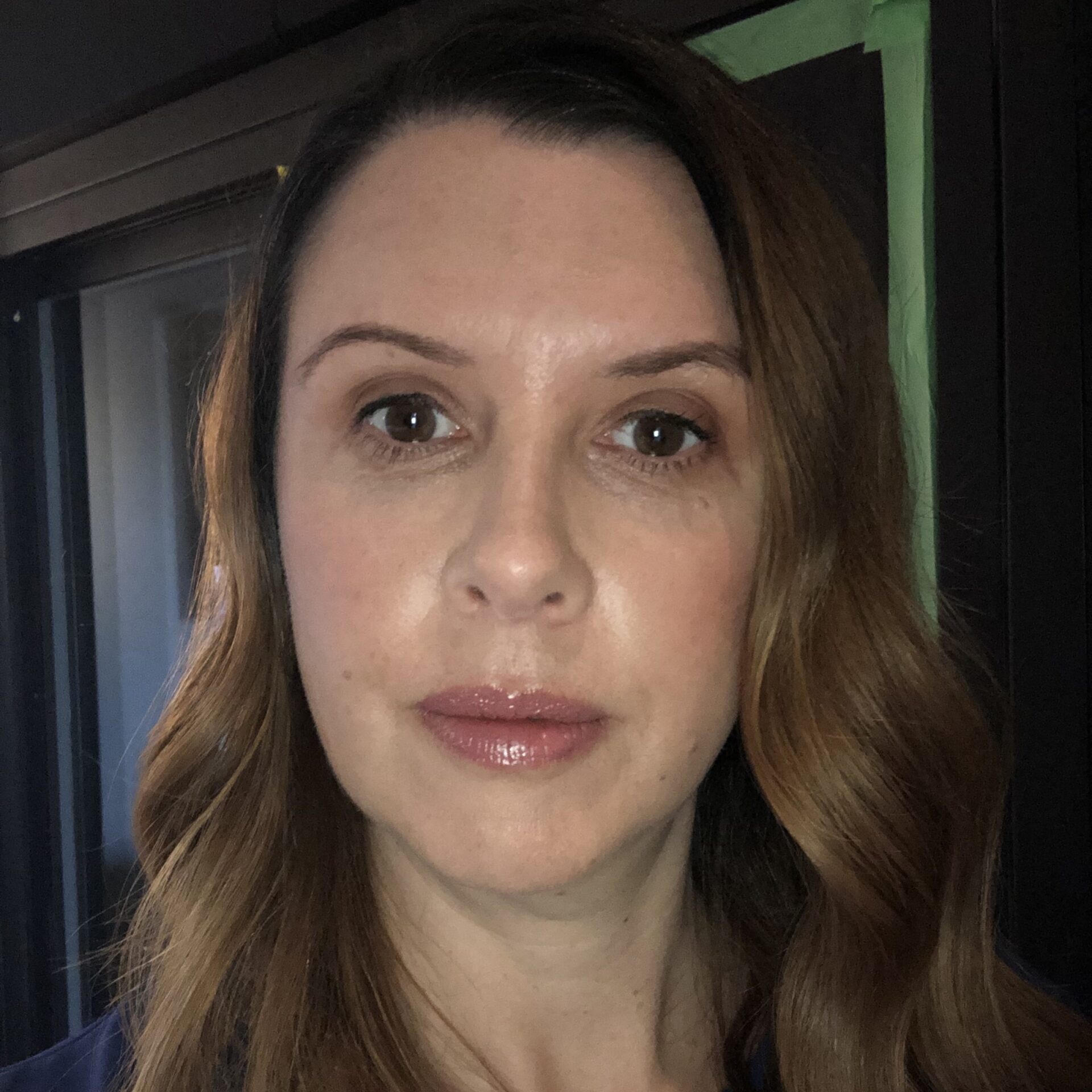 Close-up headshot of a woman with long brown hair, natural makeup, and neutral expression indoors with dark background.