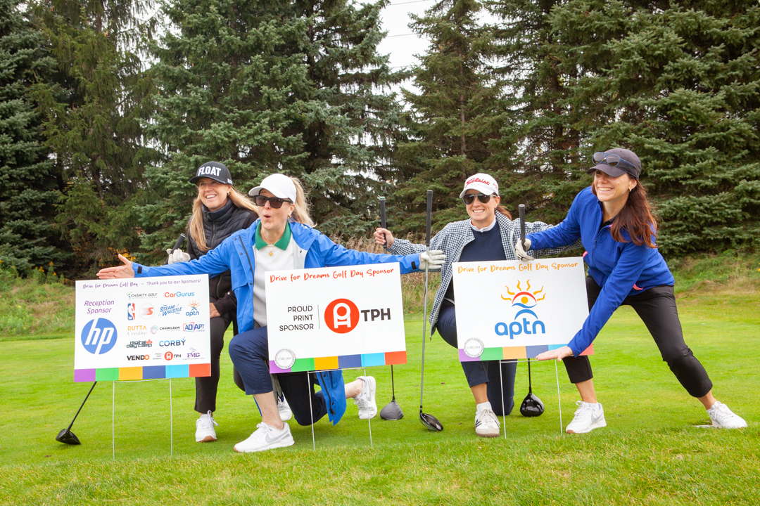 Four women pose with sponsor signs and golf clubs at the Drive for Dreams charity golf day.